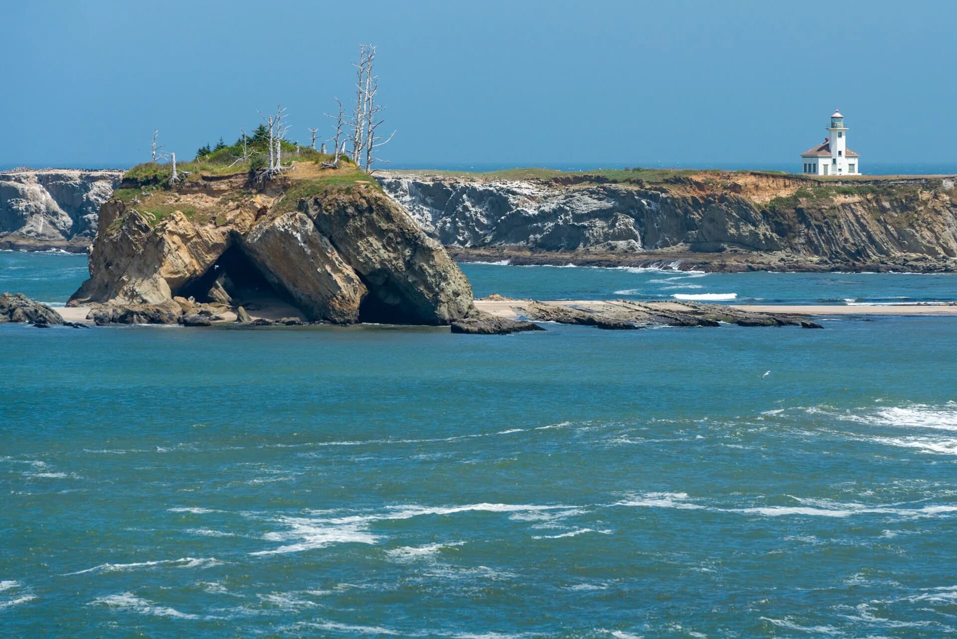 Cape Arago Lighthouse beyond rocky islands and blue coastal water.