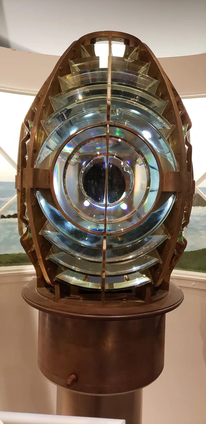 Glass Fresnel lens on a brass-colored pedestal inside a museum.