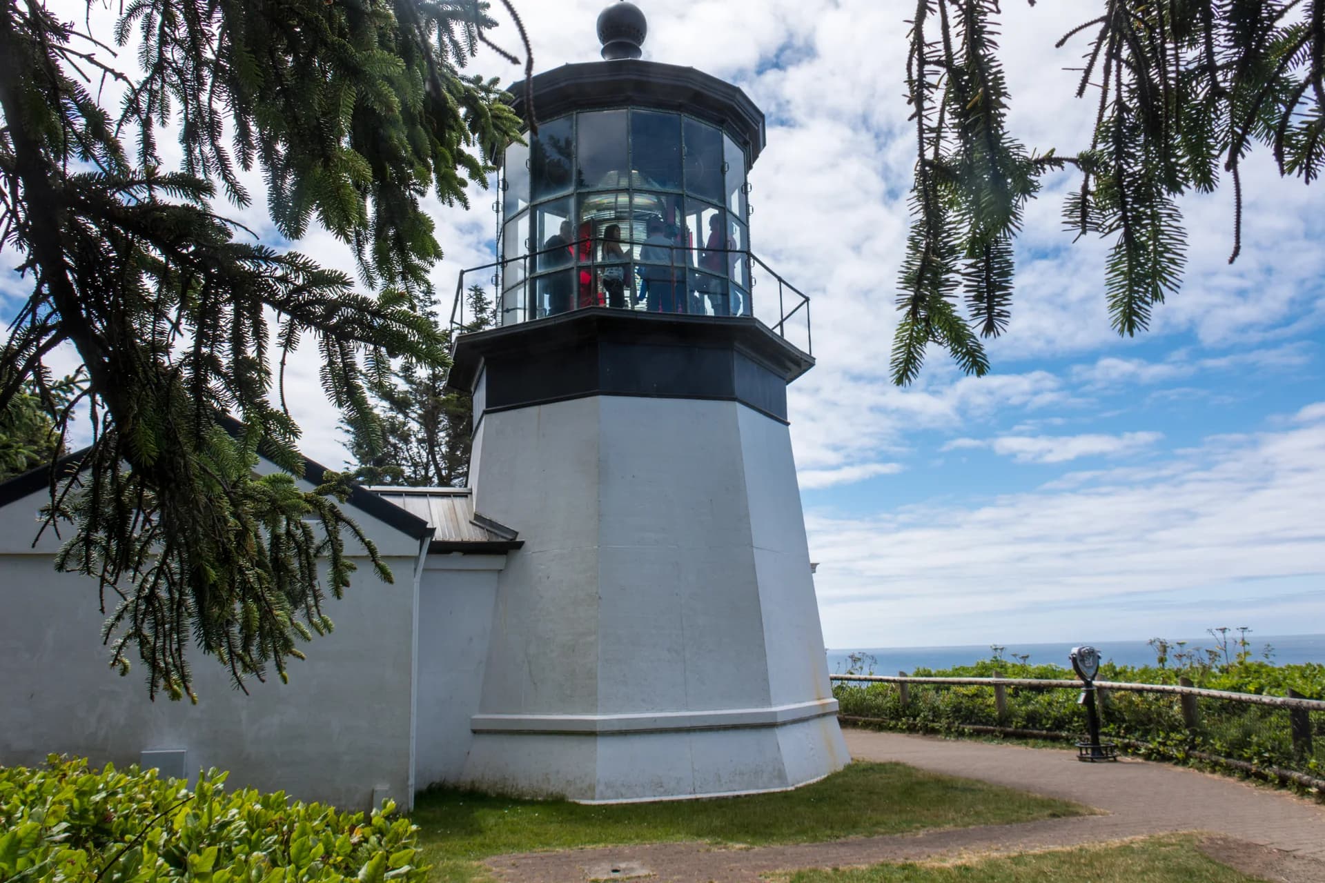 Short white Cape Meares tower beneath overhanging branches, with people visible behind its lantern glass.