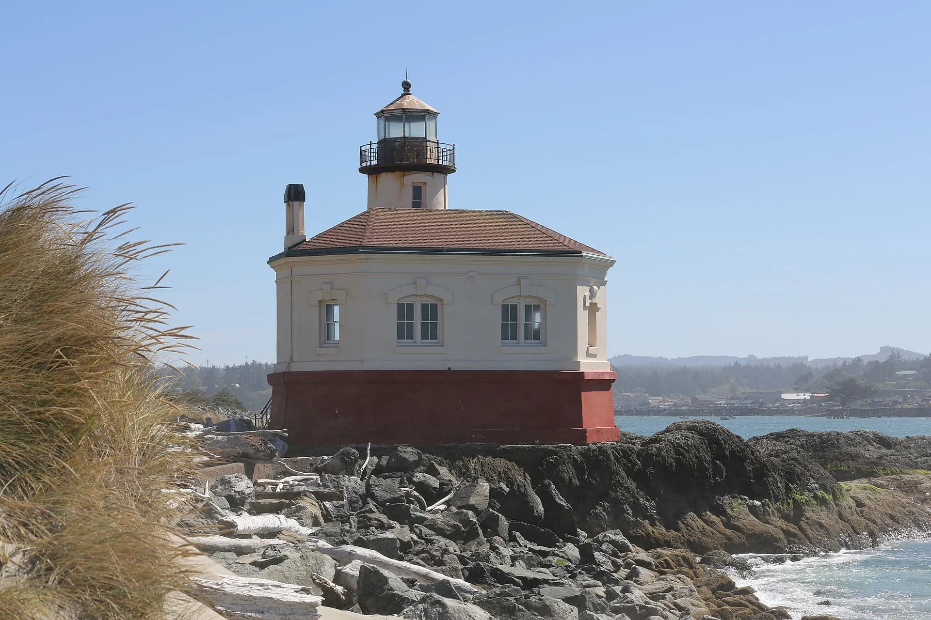 Coquille River lighthouse with a cylindrical tower behind its low building at the rocky river mouth.