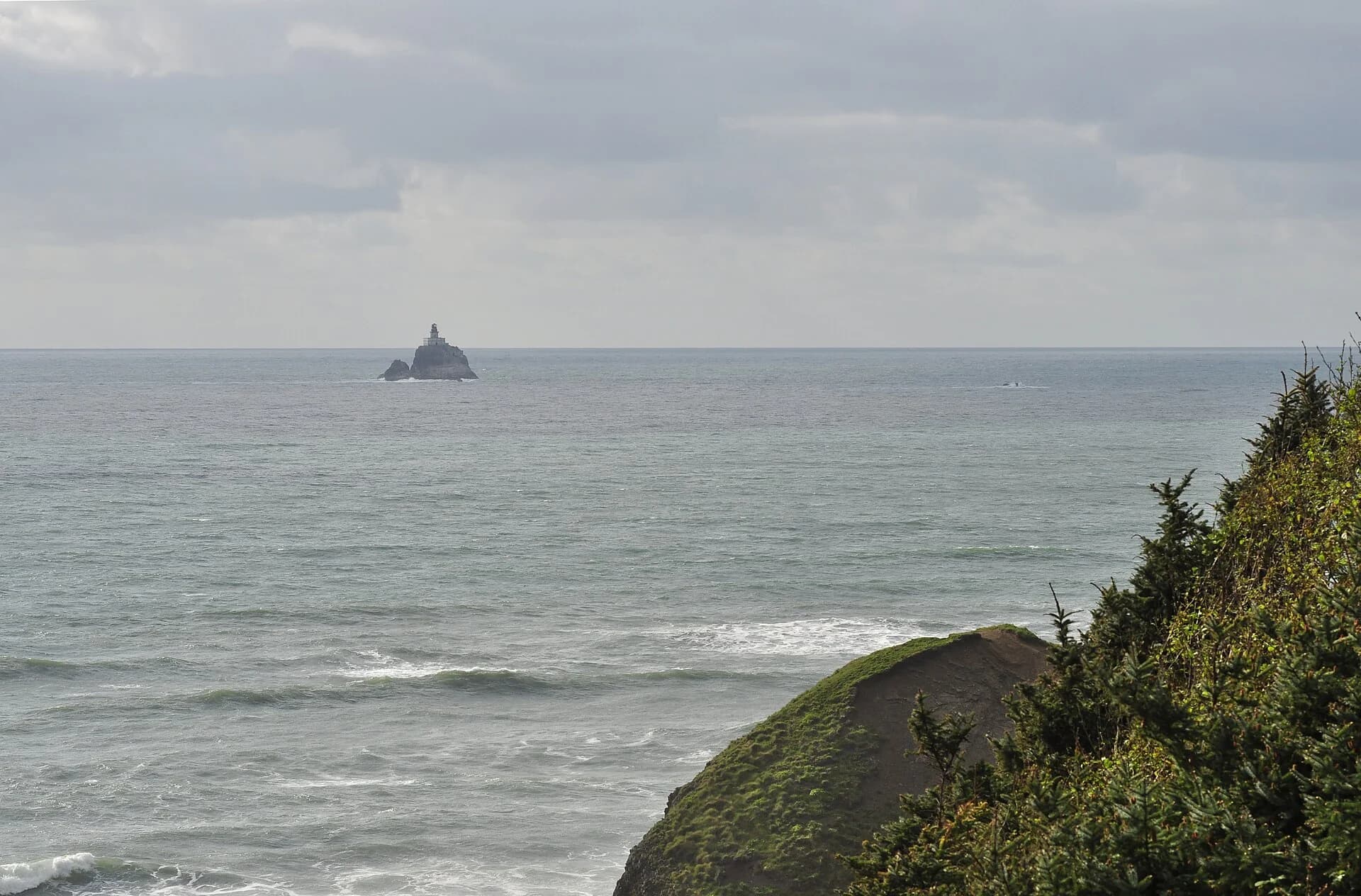 Small offshore lighthouse in a wide ocean view beyond a forested headland.