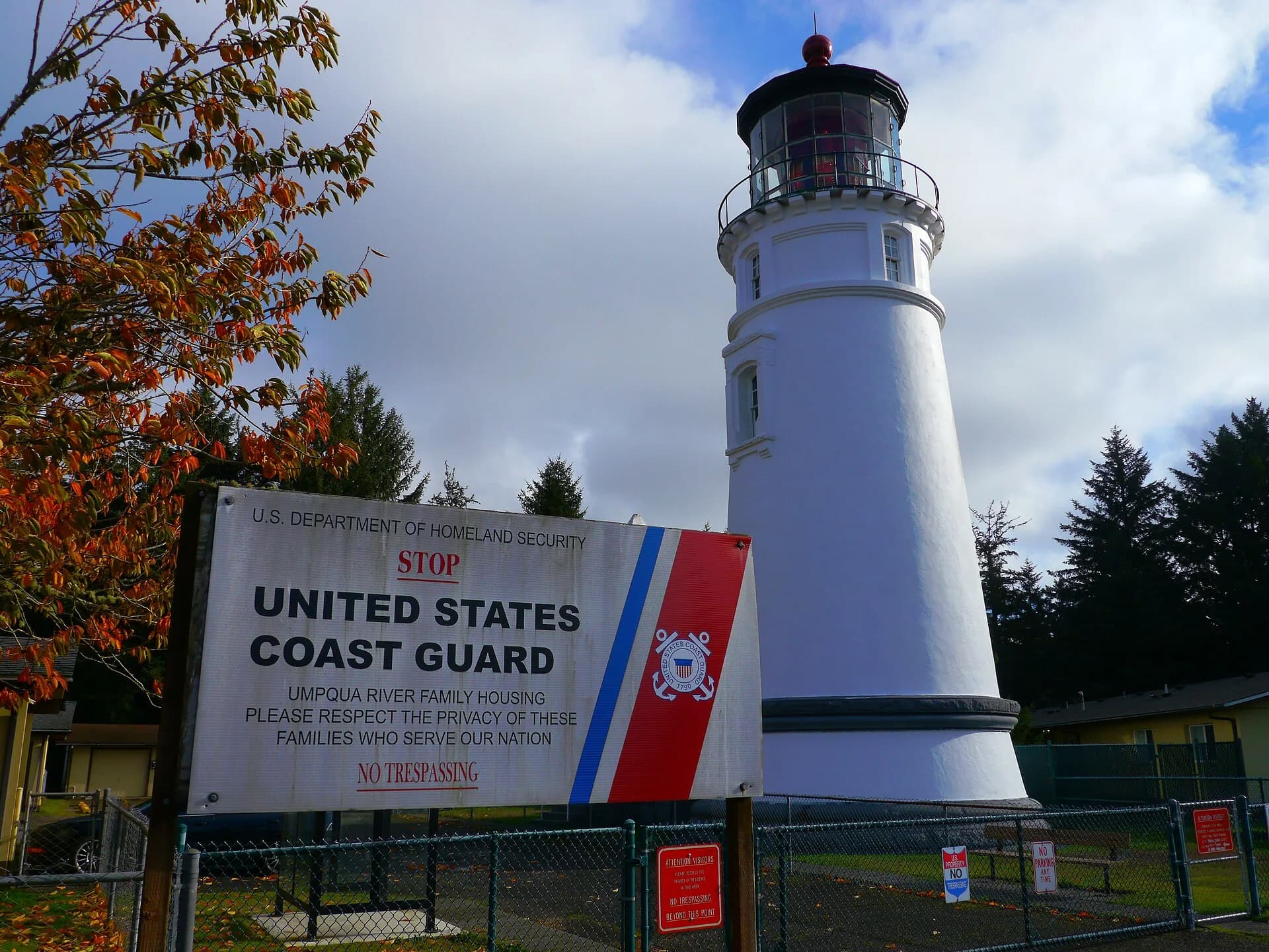 White Umpqua River lighthouse behind a Coast Guard sign and trees.