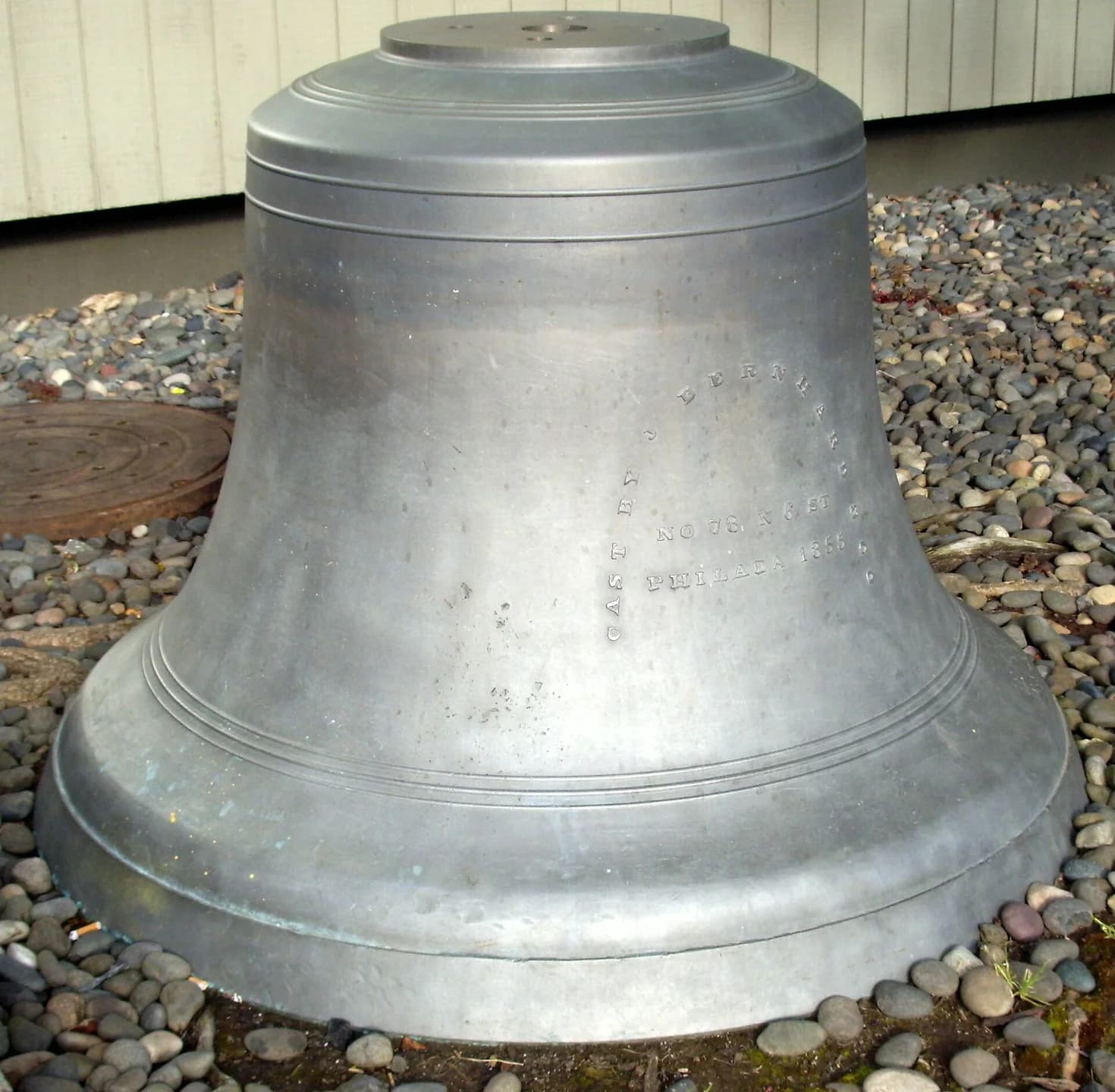 Large metal fog bell displayed over a bed of stones beside a building.